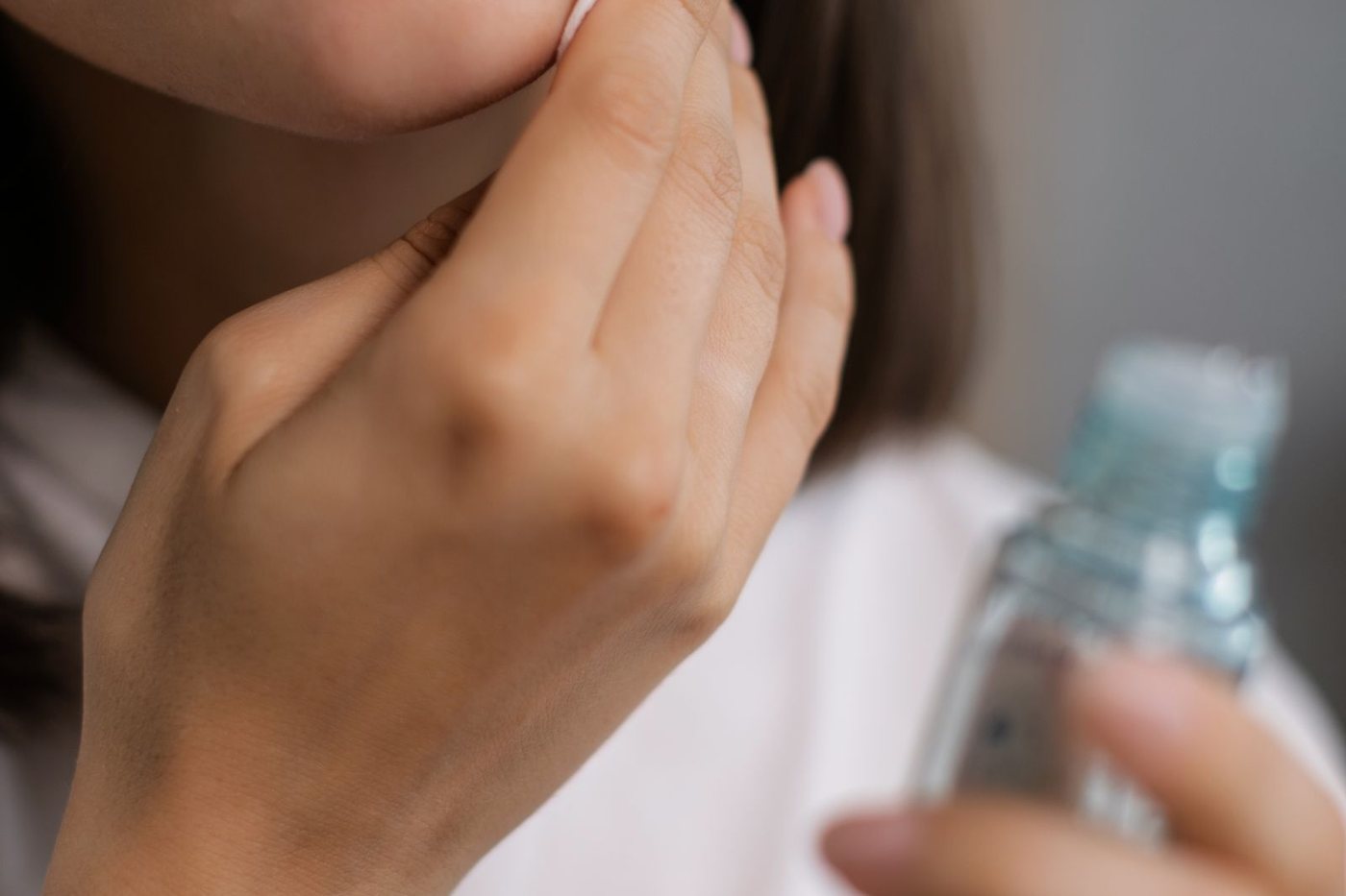 Close-up of a woman applying a clear liquid skincare product to her cheek with a cotton pad, holding an unlabelled clear bottle.