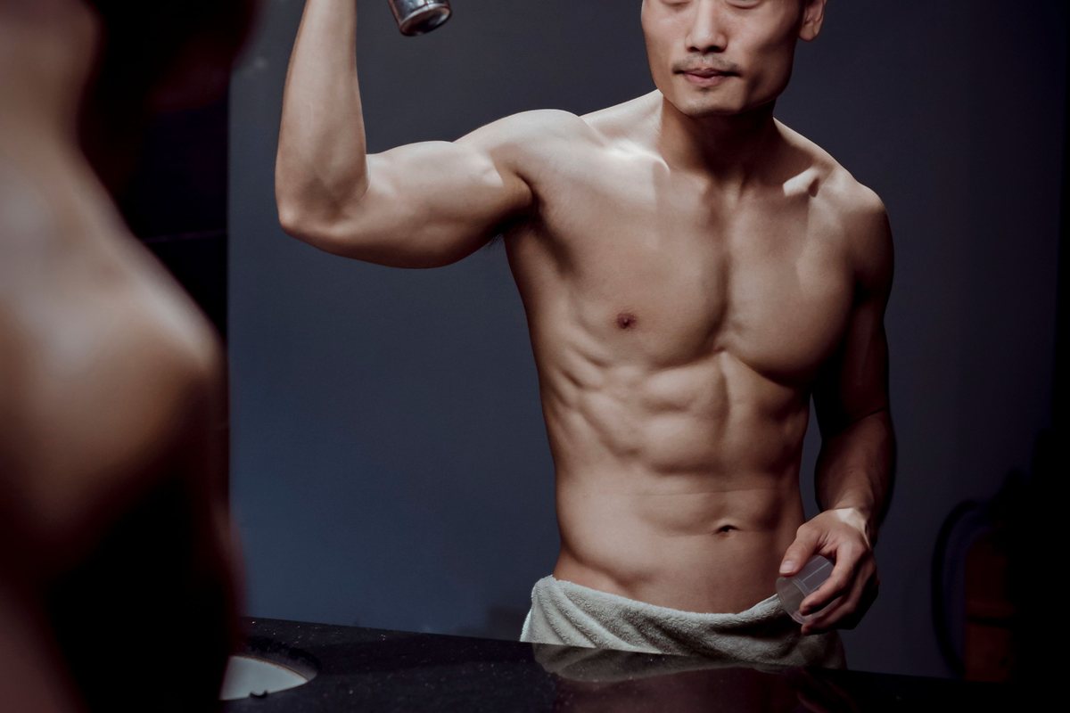 A man spraying deodorant under a raised arm in front of a bathroom mirror, with mist visible.