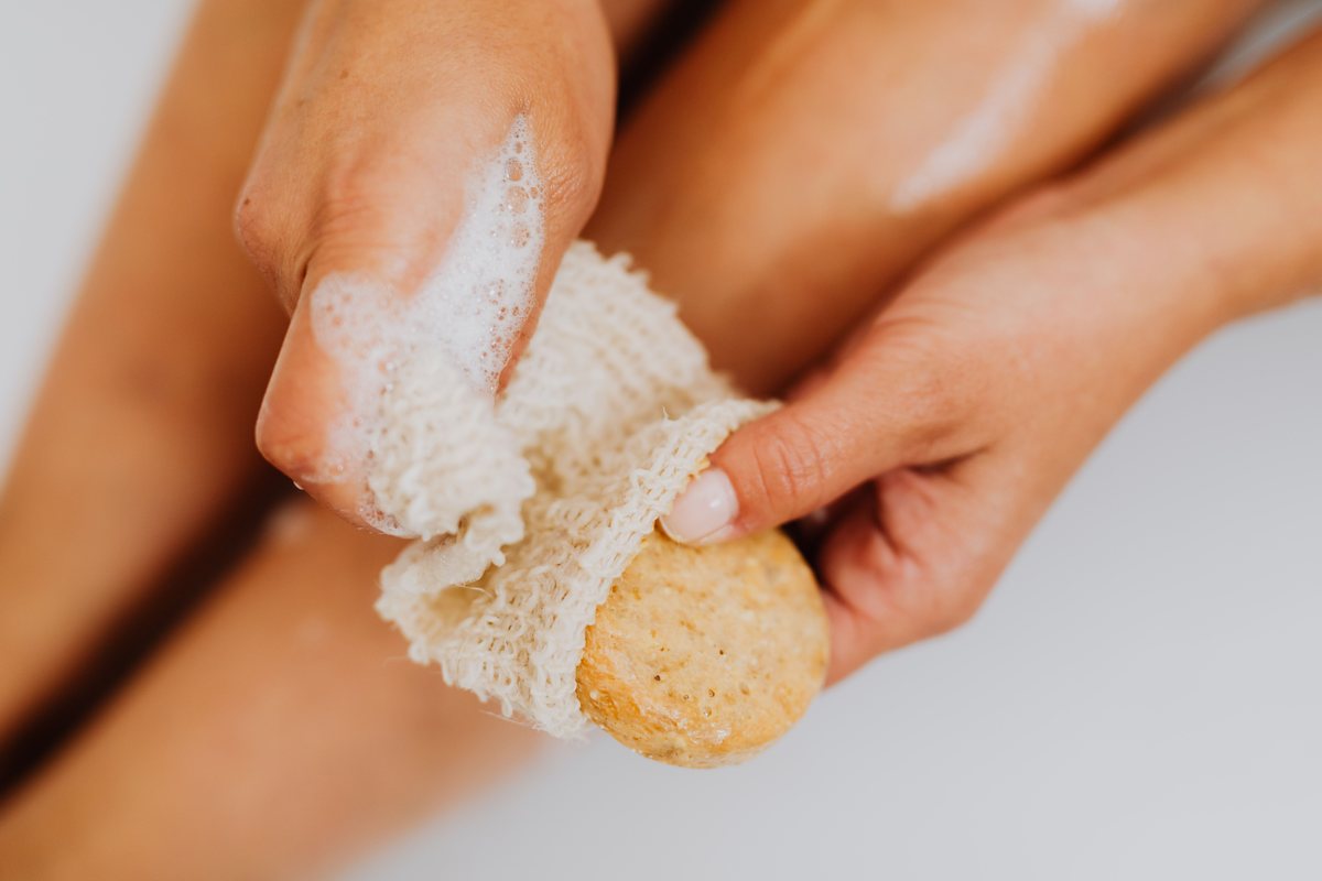 Close-up of a person's foamy hands holding a natural bath sponge, with soap lather on skin — no faces visible.