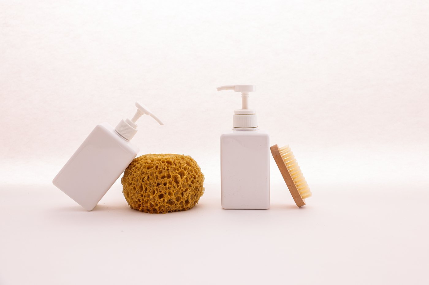 Clean white unbranded pump bottles paired with a natural sponge and wooden brush on a white background, styled as minimalist bathroom essentials.